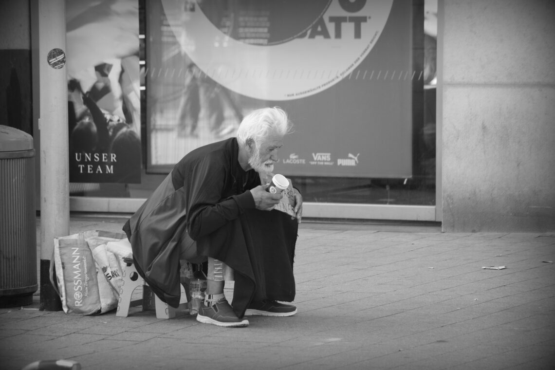 A white-haired man sitting on a plastic stool in front of two shopping bags holding a sign and a small paper cup, begging.