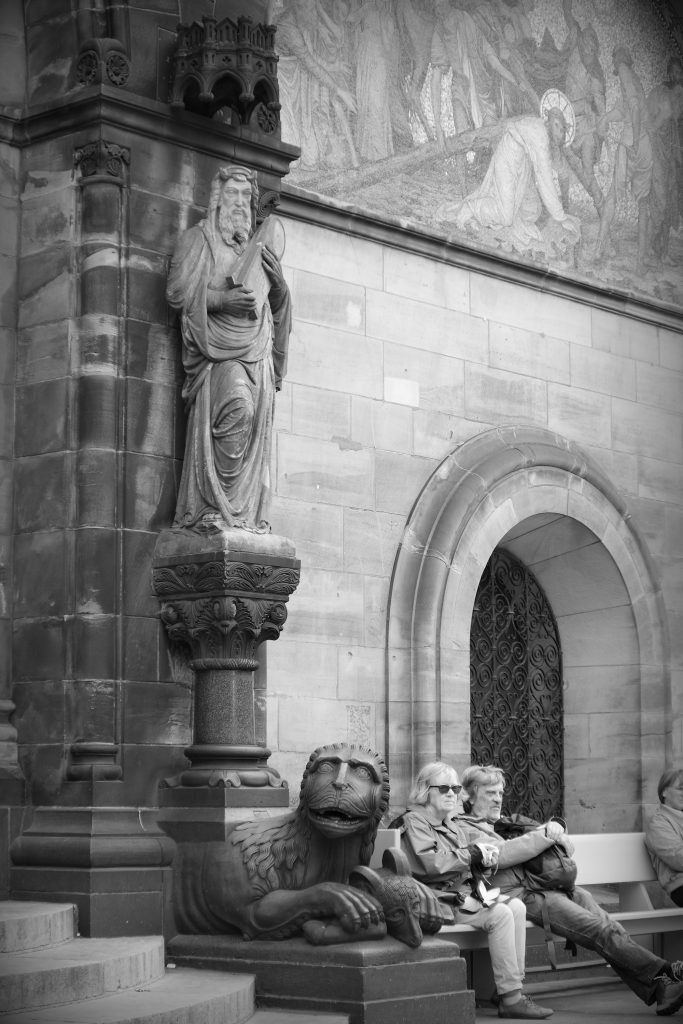 Mythical lion cradling a goat between its paws in front of the Dom in Bremen, looking toward the camera, beneath the statue of a man draped in flowing robes and carrying two stone tablets.