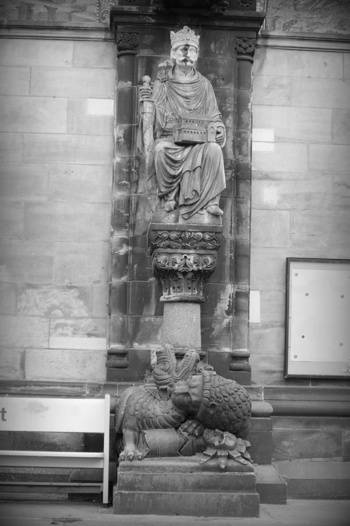 Mythical lion biting ther neck of a dragon beneath the statue of a king or bishop cradling the model of a church building, holding a sword.