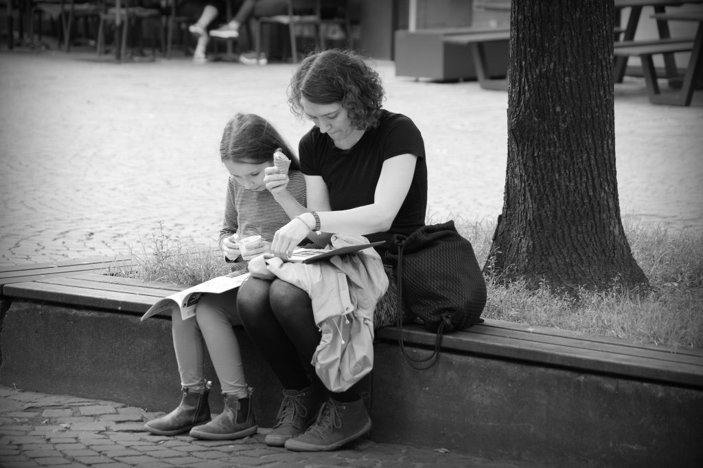 Mother and daughter in Bremen sharing time together: both eating ice cream, both reading, on a park bench under a tree.
