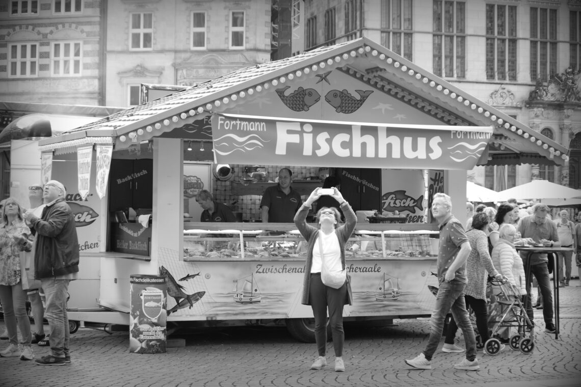 A fish stand called Fischhus in the Marktplatz, Bremen, with several people standing around looking up toward the sky where trapeze artists are performing.