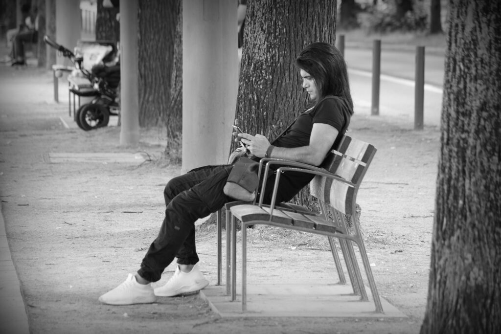 A long, dark-haired man sitting on a park bench overlooking the lake in Hamburg, checking his mobile.