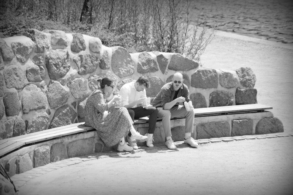 A group of three people sitting on a long wooden bank in front of a stone wall, alongside the lake in Hamburg, eating from Asian takeaway containers with chopsticks.