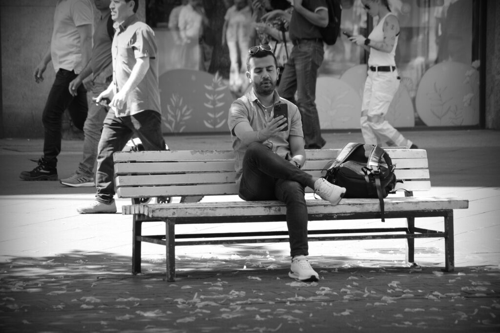 A young man on a street bench holding a smartphone out at arm's length as if taking a selfie.