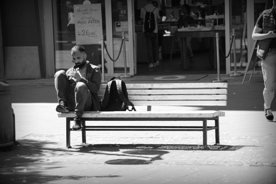 A single man on a street bench, drawing his feet up off the ground as he looks at his smart phone.