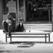 A single man on a street bench, drawing his feet up off the ground as he looks at his smart phone.
