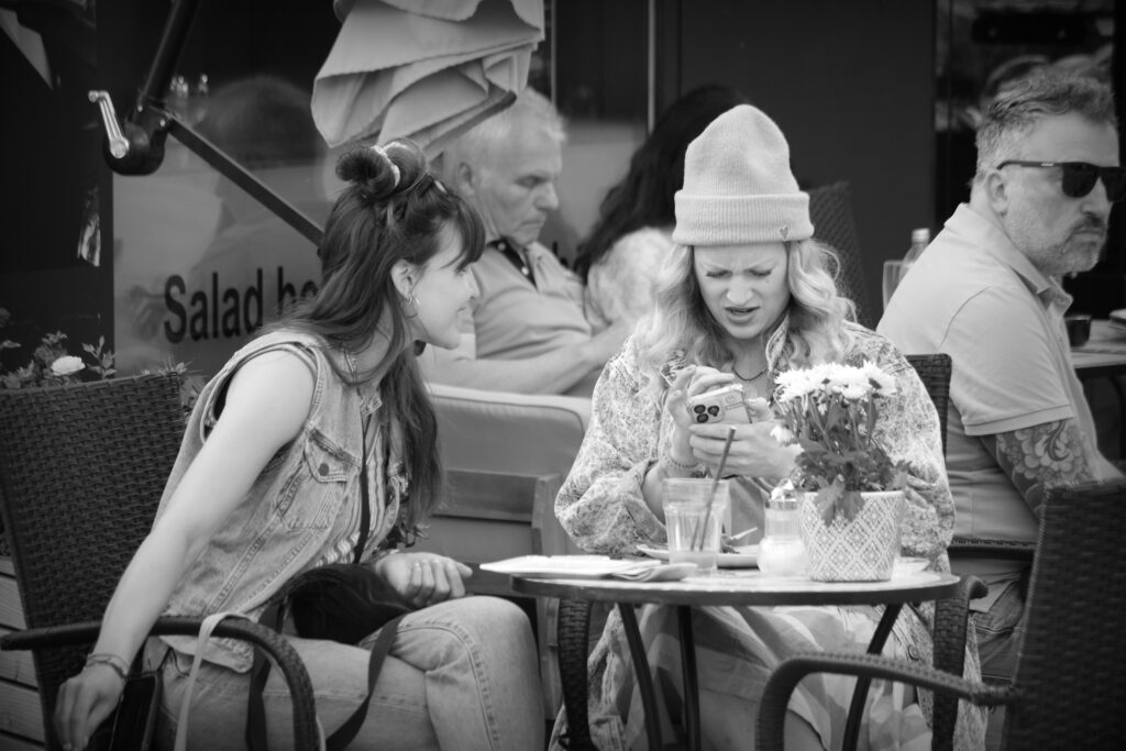 Two women at a coffee house table looking confused over a smart phone.