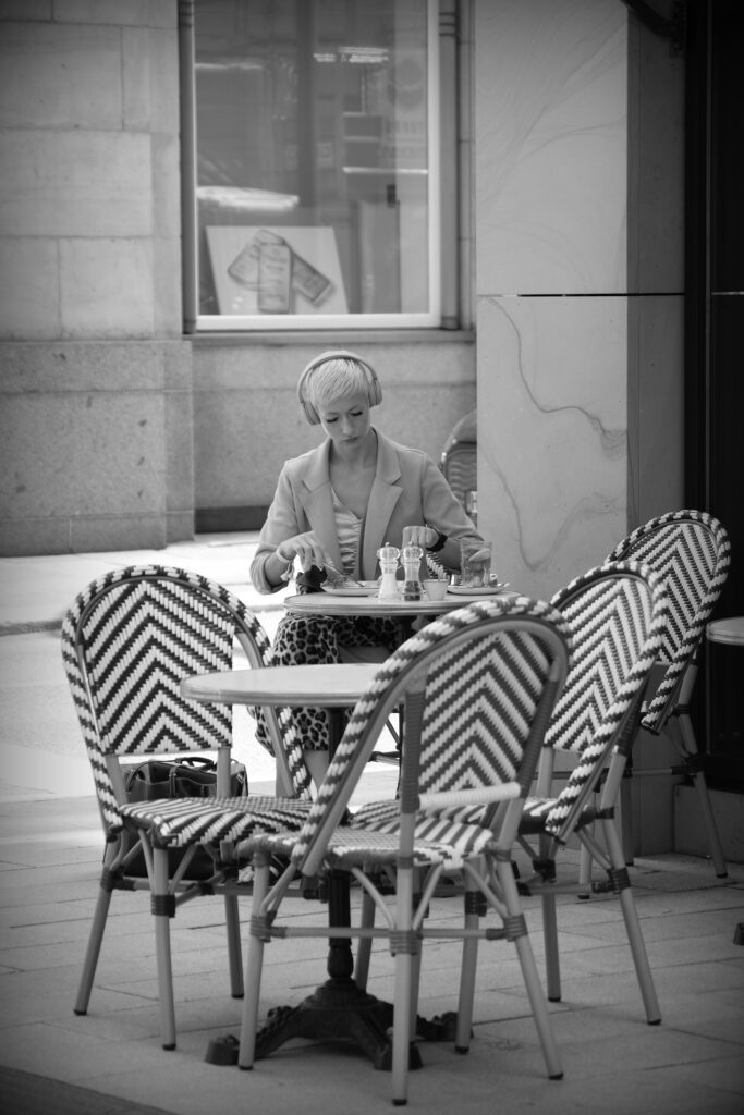 A woman wearing headphones, eating at a roadside table in front of a restaurant in Hamburg.