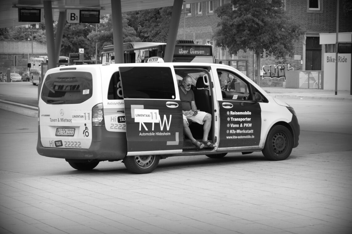 A taxi driver in shorts, sitting with his feet hanging out of the back passenger door of his vehicle in Hildesheim.