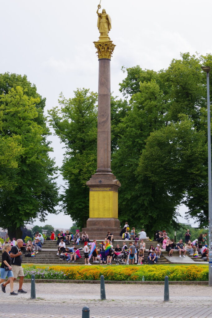 Across from the main area for CSD in Schwerin, marchers gather after the parade to rest under a memorial statue to war dead.