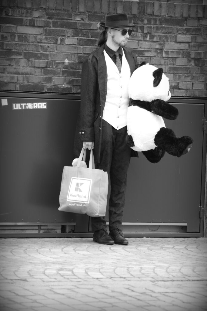 A man with a panda bear, dressed in a long jacket, white waistcoat, hat and sunglasses, with a Kaufland shopping bag, in Bremerhaven.