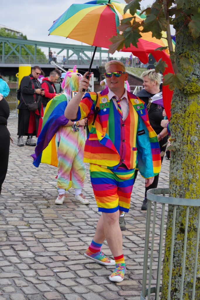 A man holding a rainbow umbrella, wearing a rainbow suit with waistcoat in Bremerhaven