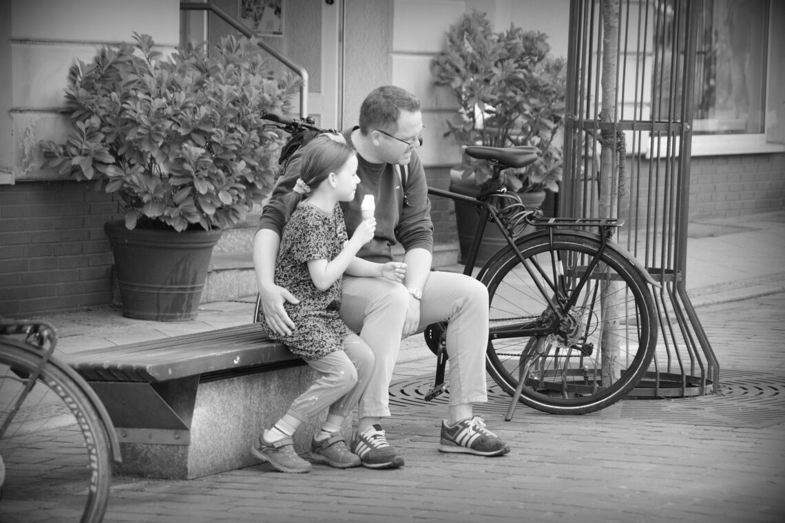 Father and daughter talking about a small dog which came near, the daughter eating a soft ice cream from a cone, the father with his arm around her. Schwerin.