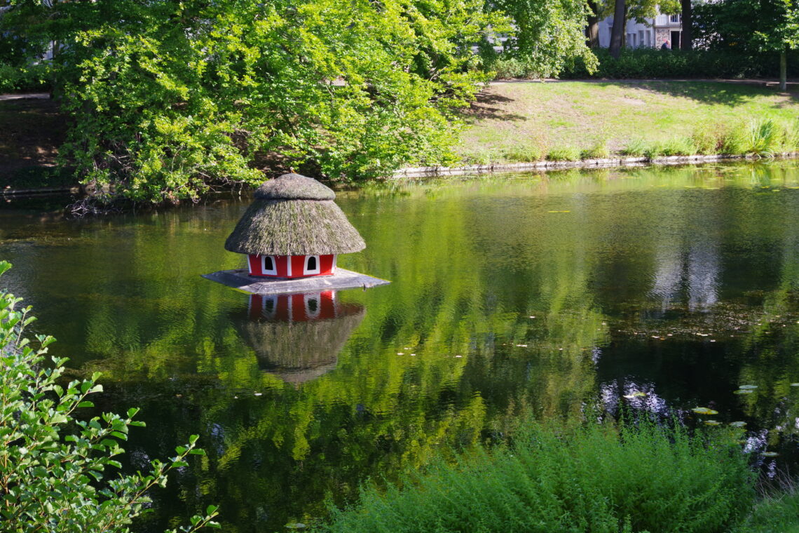 A small red, thatched-roofed duck house with white-framed open doors, in a green-colored lake surrounded by a grassy bank and summer trees in Bremen, Germany.