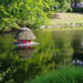 A small red, thatched-roofed duck house with white-framed open doors, in a green-colored lake surrounded by a grassy bank and summer trees in Bremen, Germany.