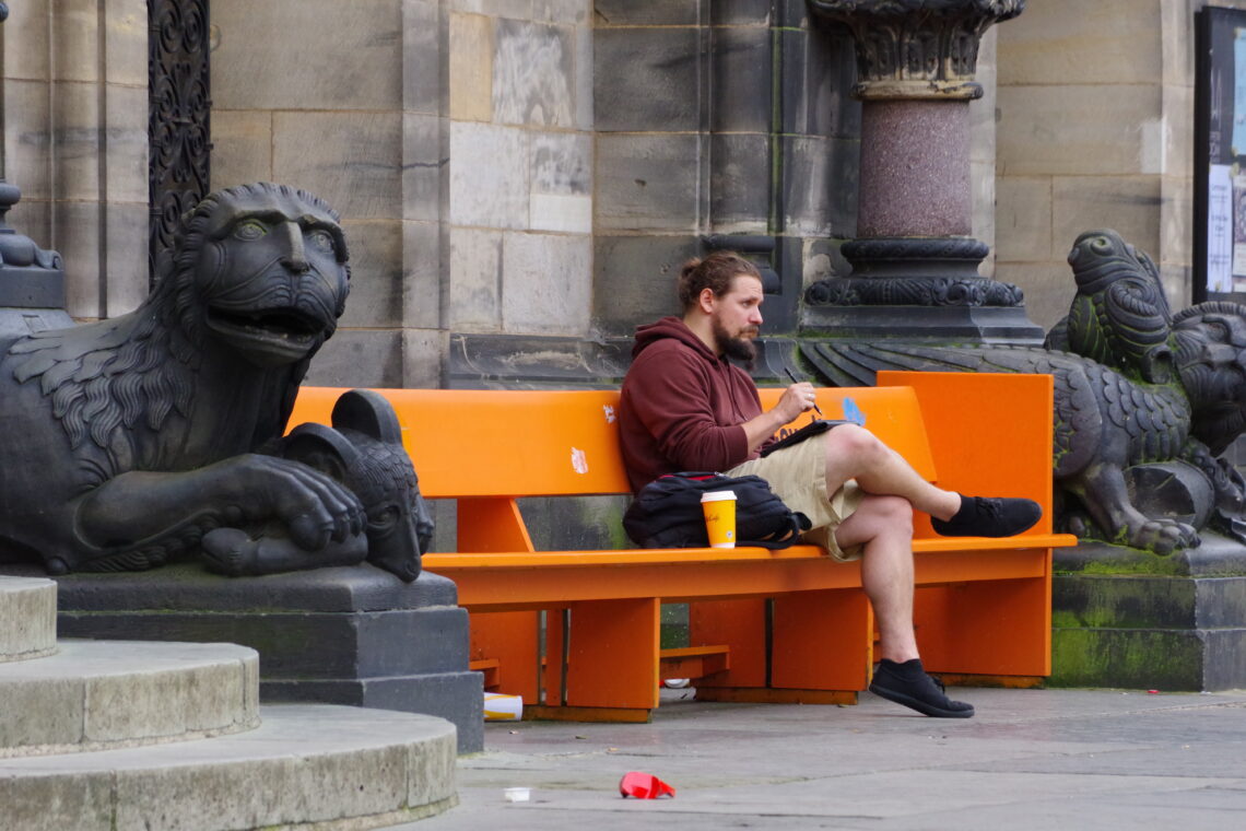 A bearded man wearing a hoodie and shorts, writing on an electronic device, sitting with a takeaway coffee on an orange "anti-violence" bench between gargoyles in Bremen.