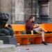 A bearded man wearing a hoodie and shorts, writing on an electronic device, sitting with a takeaway coffee on an orange "anti-violence" bench between gargoyles in Bremen.