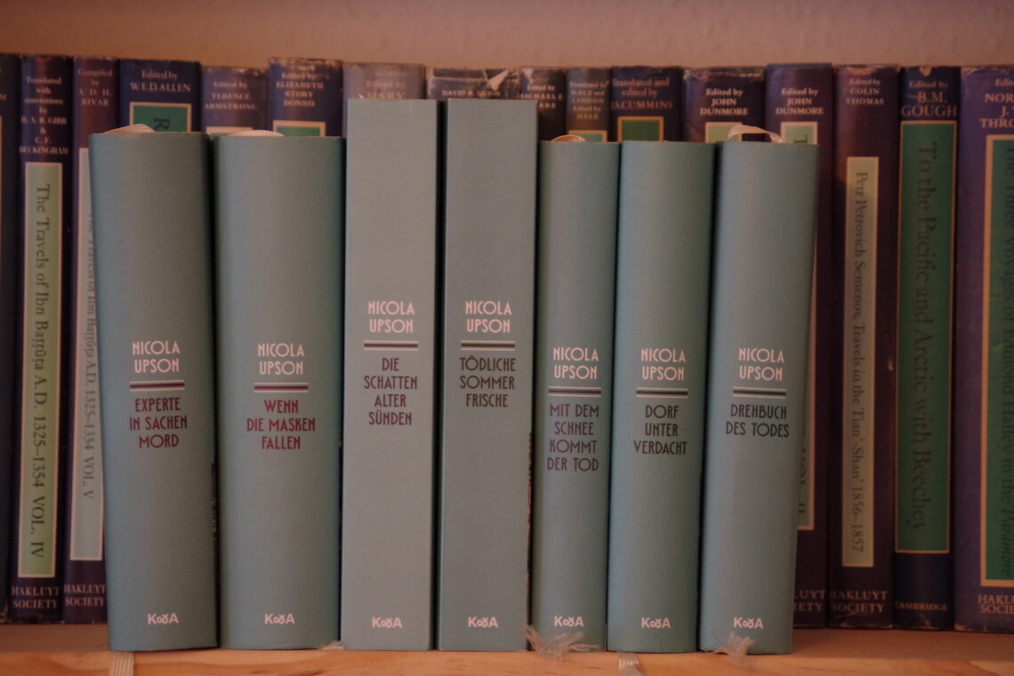 A non-symmetrical row of seven books, two of which are paperback, with blue-grey covers, titled on the side in white and black.