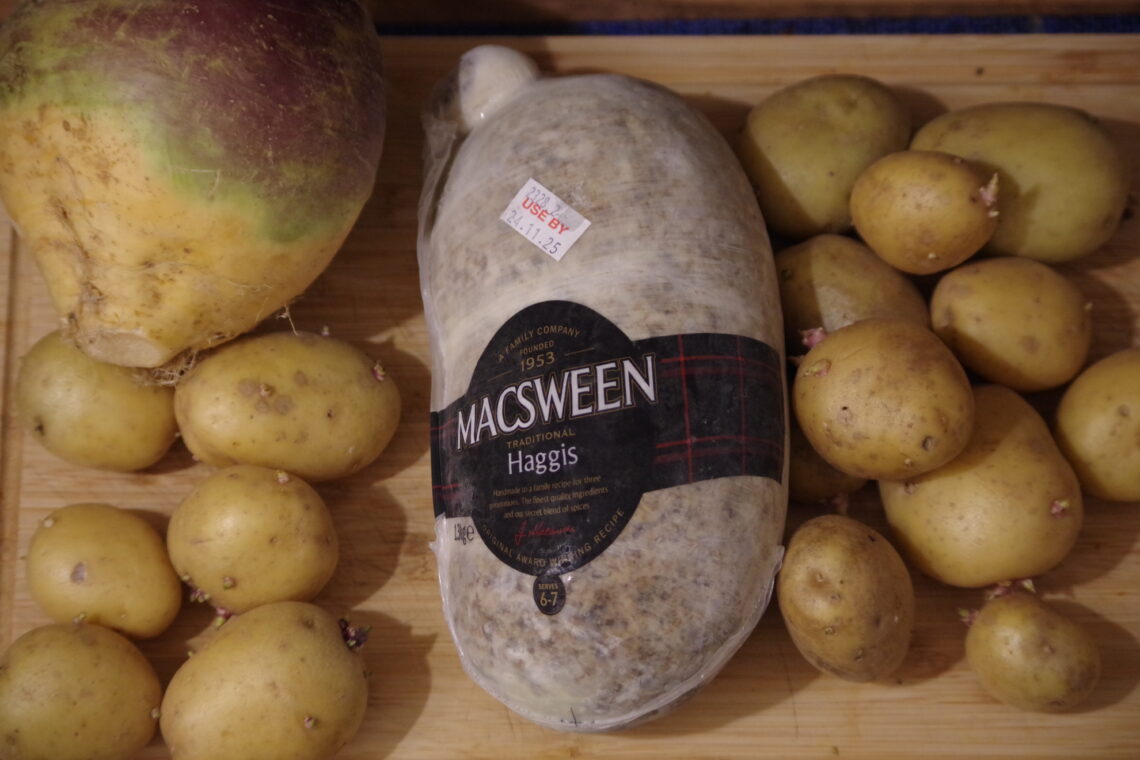 A wooden cutting board with a turnip, some potatoes and a wrapped, frozen MacSween Haggis.