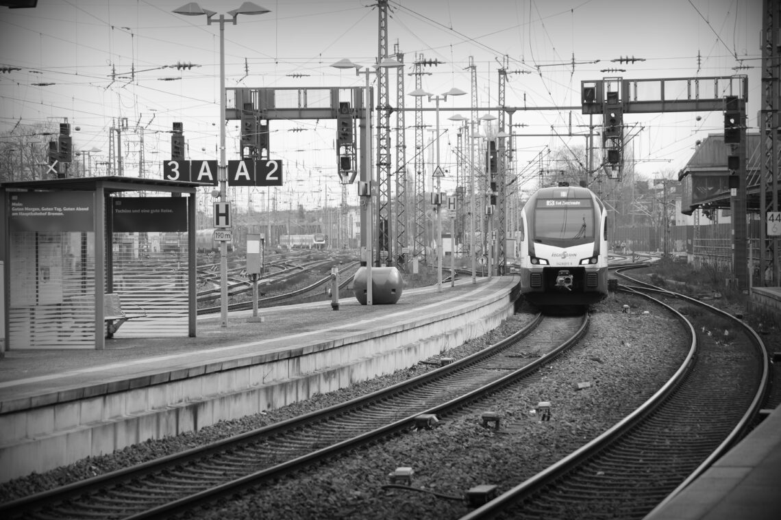 A black and white photograph of a main train station, with railway lines, masts wires and various lights forming a barrier to seeing further. In the distance there are several parked trains, and a single train is pulling slowly into the station alongside a concrete platform.