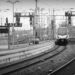 A black and white photograph of a main train station, with railway lines, masts wires and various lights forming a barrier to seeing further. In the distance there are several parked trains, and a single train is pulling slowly into the station alongside a concrete platform.