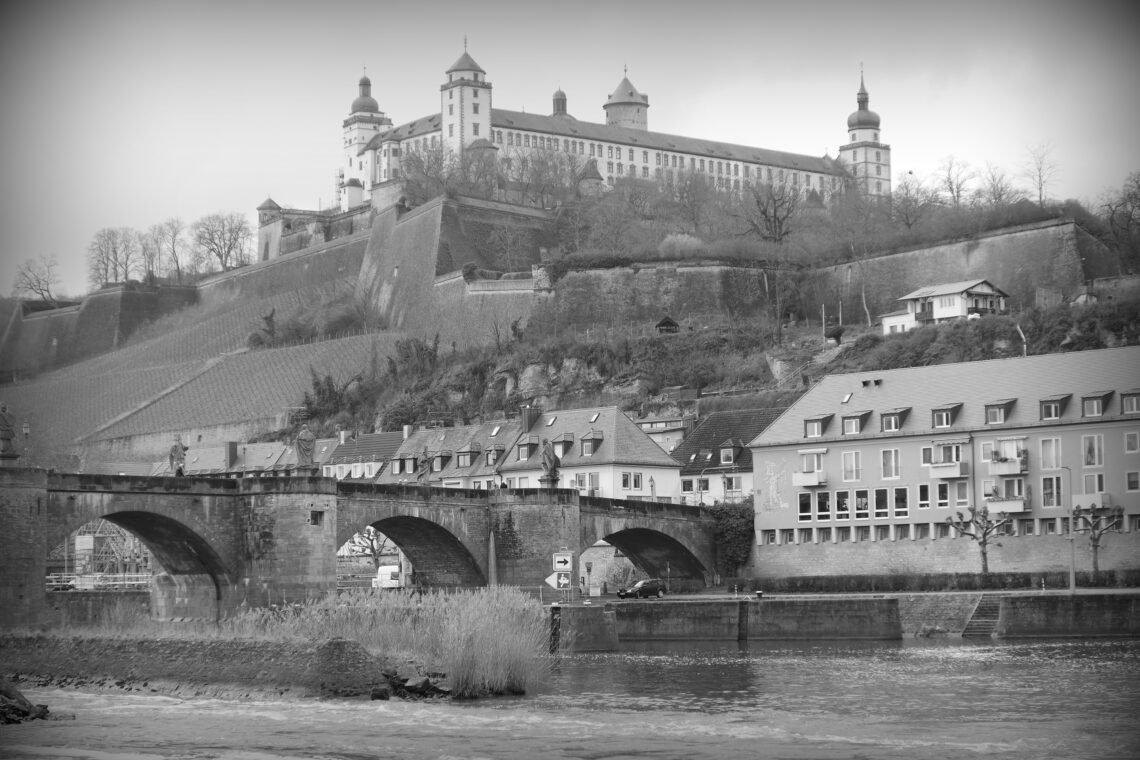 Black and white photograph of a castle with various turrets high on a hill, with a steep drop down over vineyards to houses and a brick bridge across a raging river. Würzburg.