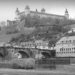 Black and white photograph of a castle with various turrets high on a hill, with a steep drop down over vineyards to houses and a brick bridge across a raging river. Würzburg.