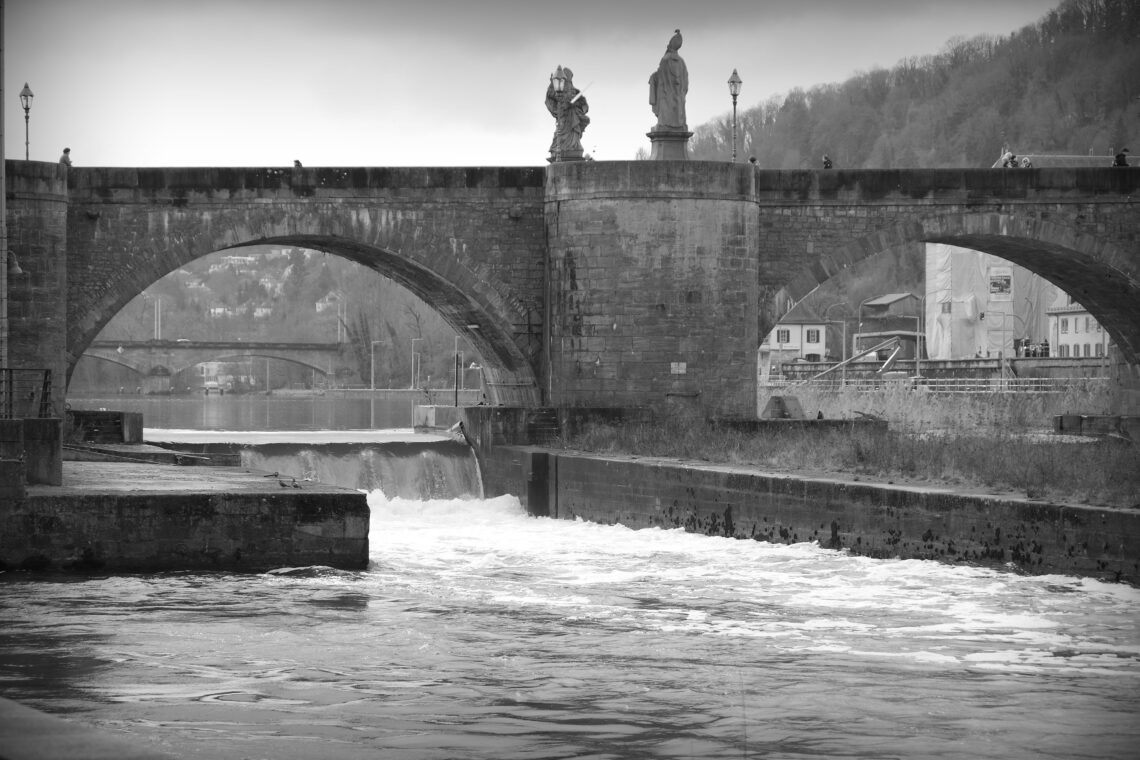 A black and white photograph through one of two arches of a bridge over the river Main in Germany. In the background there are the arches of another stone bridge, in the foreground a rush of water downriver.