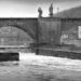 A black and white photograph through one of two arches of a bridge over the river Main in Germany. In the background there are the arches of another stone bridge, in the foreground a rush of water downriver.