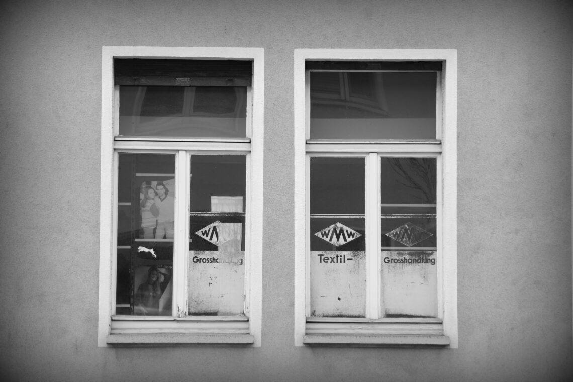 A black and white photograph of two double wood-frame windows, partially covered with white paint advertising a former textile handler. From a side street in Würzburg, Germany.