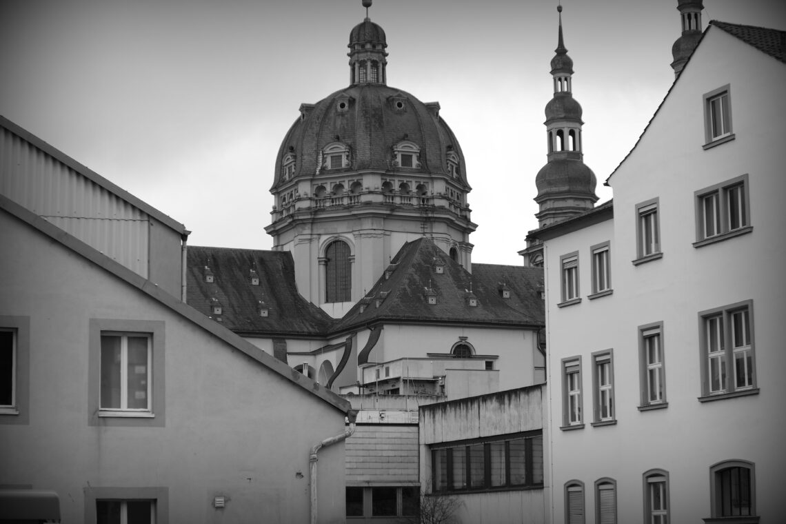 A black and white view of a religious building seen between the walls and roofs of other buildings in Würzburg.