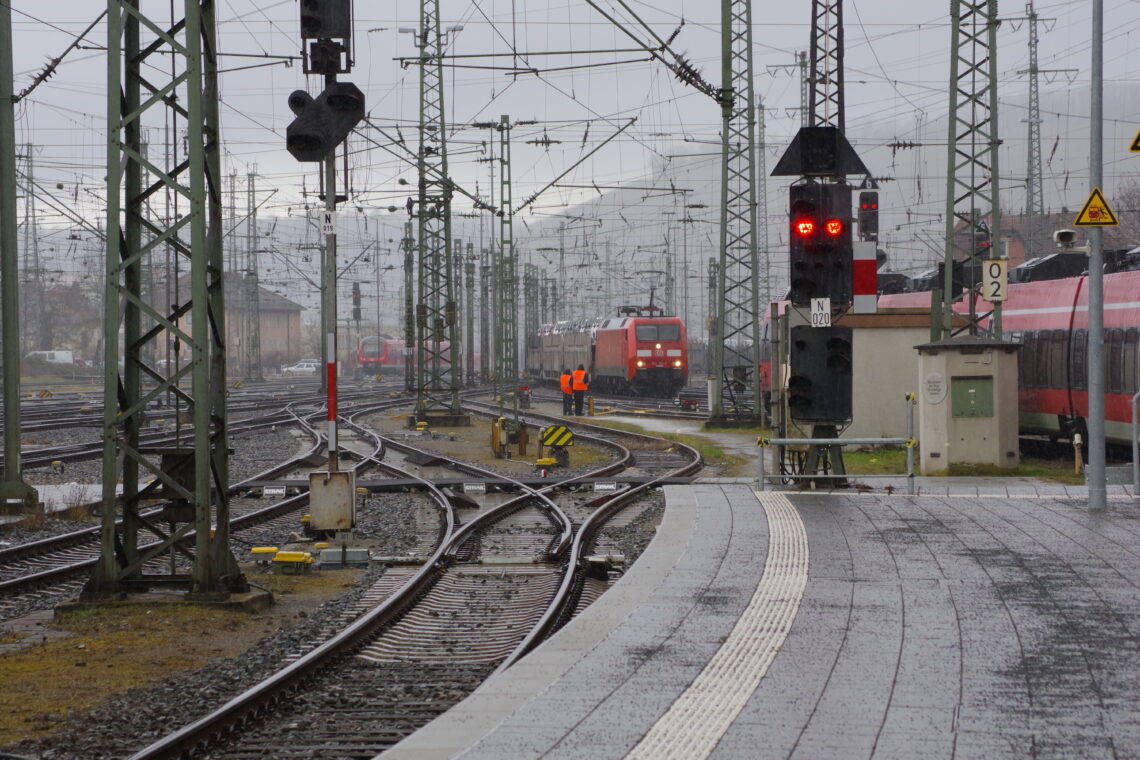 A colour photograph of winding railway tracks leading into a railway station. There is a parked train in the distance, and another strain approaches, passing two workers with high visibility vests. The scene is filled with steel supports and columns and electrical wires.