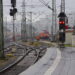 A colour photograph of winding railway tracks leading into a railway station. There is a parked train in the distance, and another strain approaches, passing two workers with high visibility vests. The scene is filled with steel supports and columns and electrical wires.