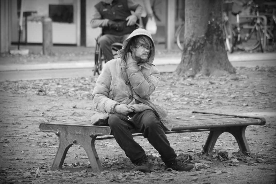 A homeless person wearing a dirty parka-style coat with the hood over his head, sitting on a park bench in Bremen. Copyright Urban Camera 2025.