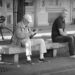 An older person sitting on a concrete bench in the shade, looking at his mobile phone, being watched by a second person. Copyright Urban Camera 2024