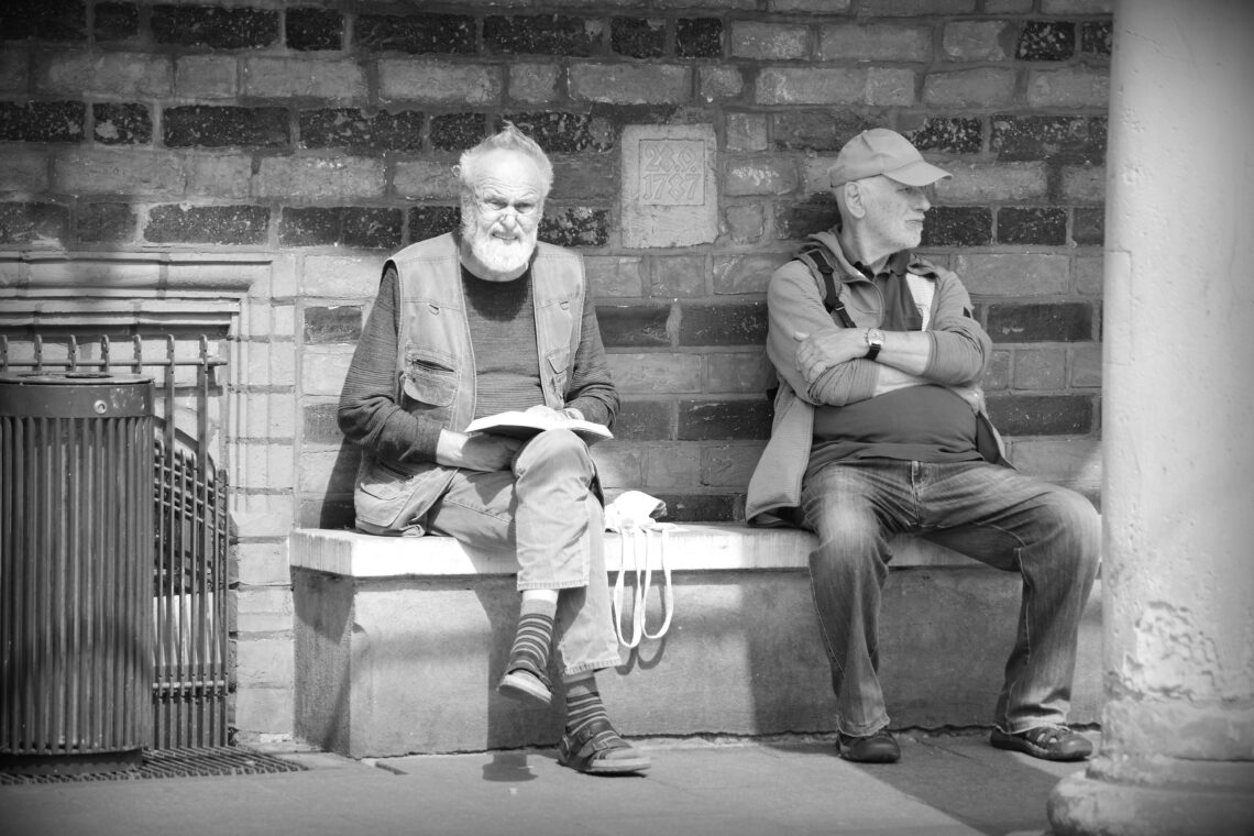 Monochrome image of two men sitting on a stone bank under the arches of the Bremen Town Hall. One has his arms folded across a generous stomach, the other is squinting into the sunshine, an open book in his hands. Taken June 2024, Copyright Urban Camera.