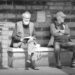 Monochrome image of two men sitting on a stone bank under the arches of the Bremen Town Hall. One has his arms folded across a generous stomach, the other is squinting into the sunshine, an open book in his hands. Taken June 2024, Copyright Urban Camera.