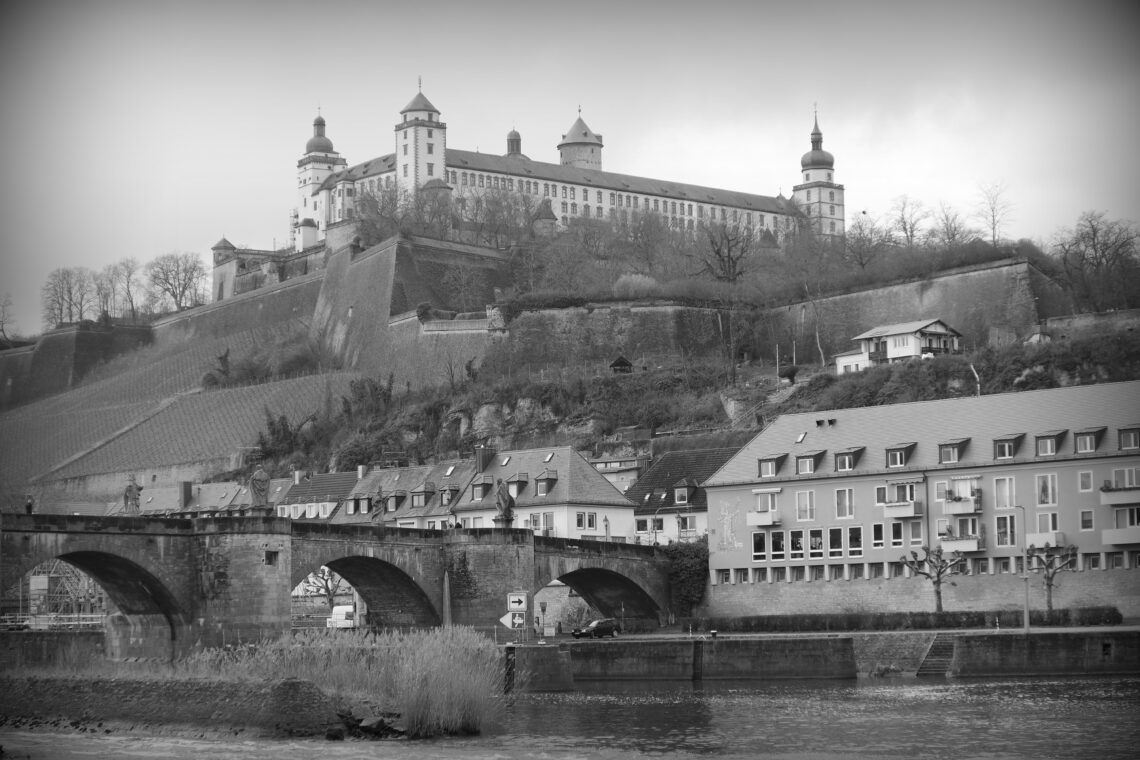 A monochrome photograph f the castle in Würzburg, Germany. The turreted building is high on a hill, beneath which are rows of houses and a stone bridge across a turbulent river. Copyright Urban Camera.