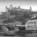 A monochrome photograph f the castle in Würzburg, Germany. The turreted building is high on a hill, beneath which are rows of houses and a stone bridge across a turbulent river. Copyright Urban Camera.