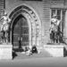 A monochrome photograph showing two people sitting on stone steps between statues of mounted, amoured knights bearing spears before a large wooden double-door with metal tracings at the Rathaus in Bremen, Germany. Copyright Urban Camera.