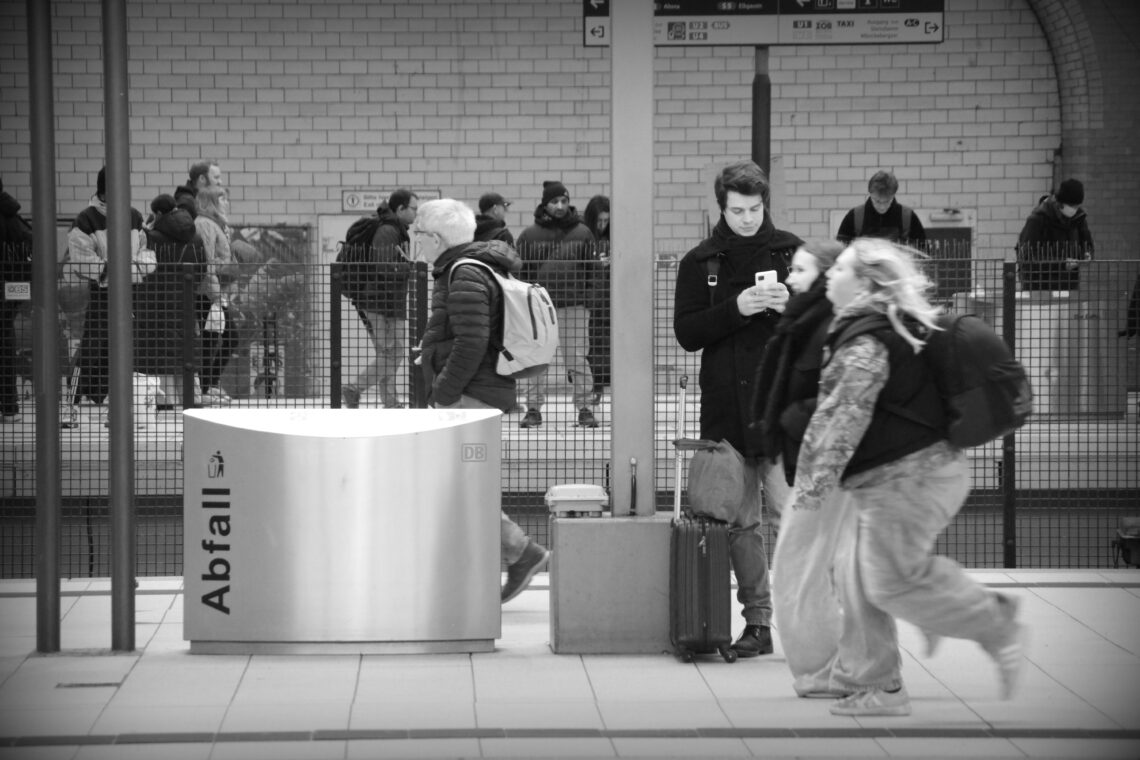 A monochrome photograph at Hamburg main railway station, showing crowds of people waiting for their train train across a platform, with two people blurred as they run for a train about to leave. Copyright Urban Camera.