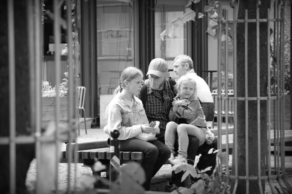 A monotone photograph of a young woman eating a hamburger with what appears to be her father berating her over something, while a smaller child sits on the man's knee. The young woman has the typical "leave me in peace" look on her face of all teenagers when a older person tells them what to do. Copyright Urban Camera.