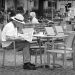 A monochrome photograph of a man wearing a white sun hat, sitting at an outside table in front of a bakery, reading a newspaper. Copyright Urban Camera.