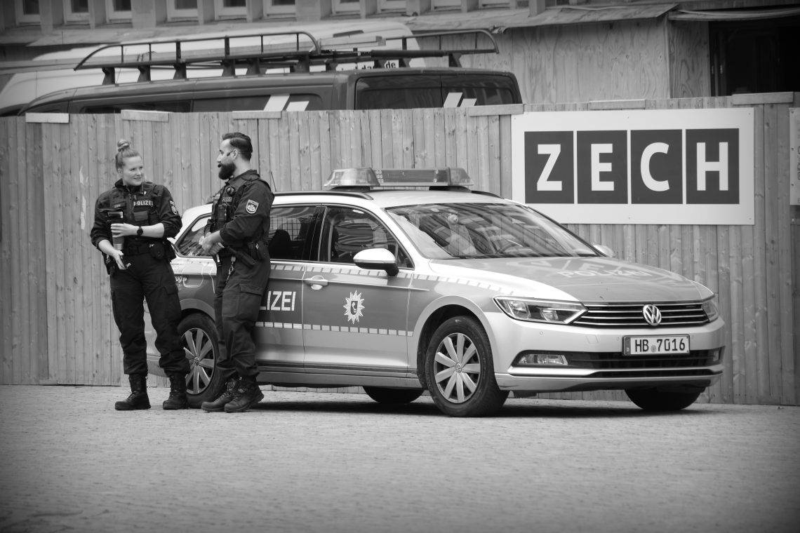 A monochrome photograph of a male and a female police officer standing in front of their vehicle by the wooden fencing of a building site in Bremen, Germany. Copyright Urban Camera.