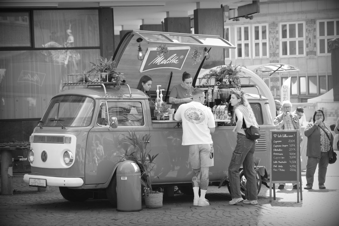 A monochrome photograph of a mobile coffee shop built into a VW traveller. The side of the VW opens upwards and customers are standing ordering drinks. Copyright Urban Camera.