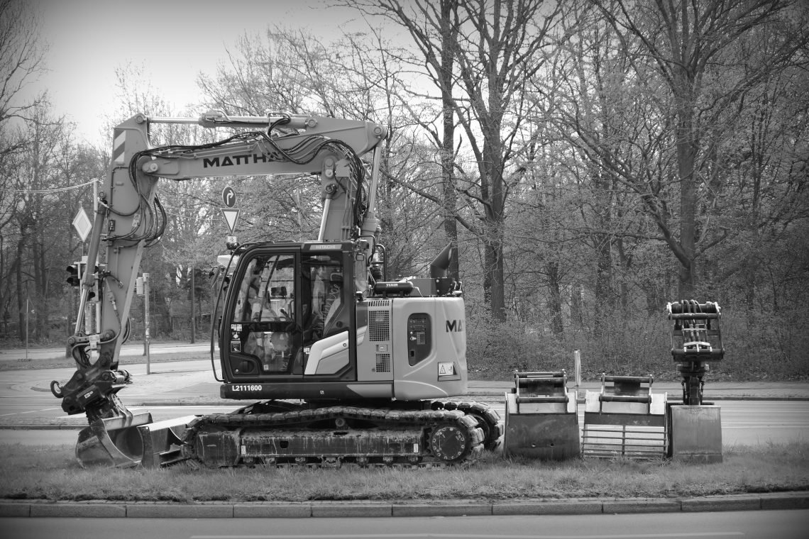 Monochrome photograph of an excavator with spare and supplementary shovels lined behind it, beside a main road, awaiting work to begin shortly. Bremen, Germany. Copyright Urban Camera.