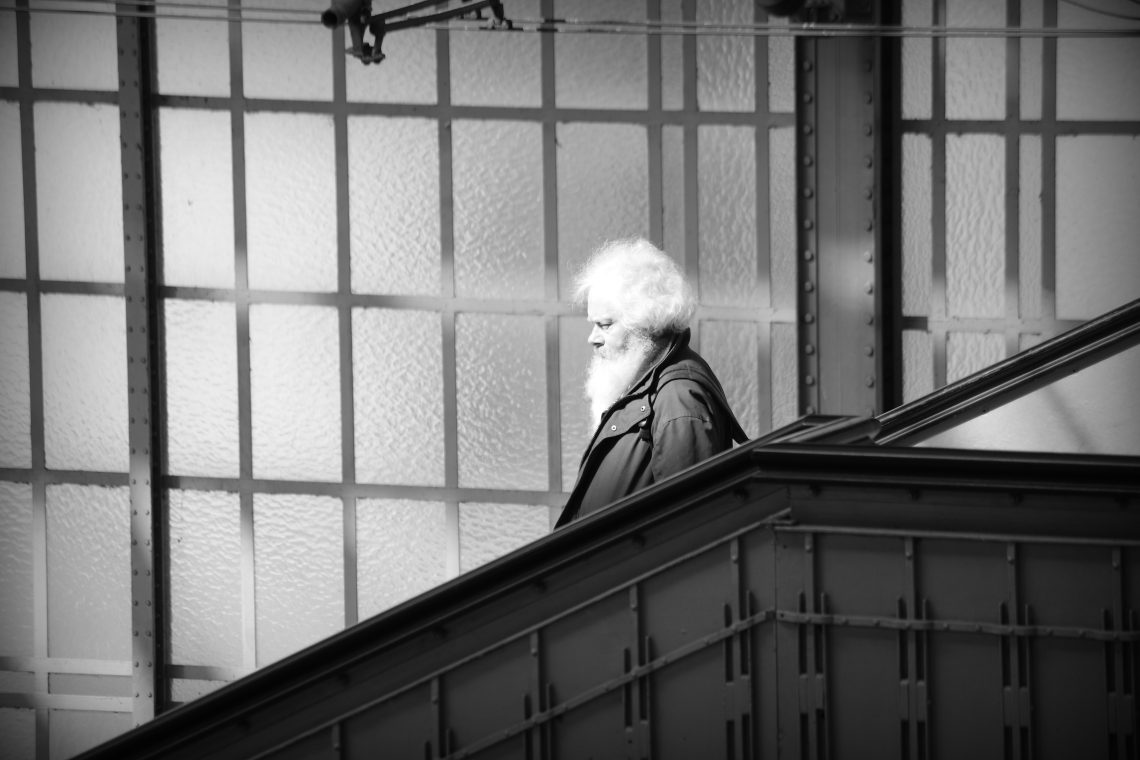 A monochrome photograph of a white haired, bearded man caught in a shaft of sunlight, descended stairs in Lübeck main railway station. Behind him is a wall of frosted glass window panes with a metal support column. Copyright Urban Camera.