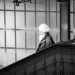 A monochrome photograph of a white haired, bearded man caught in a shaft of sunlight, descended stairs in Lübeck main railway station. Behind him is a wall of frosted glass window panes with a metal support column. Copyright Urban Camera.
