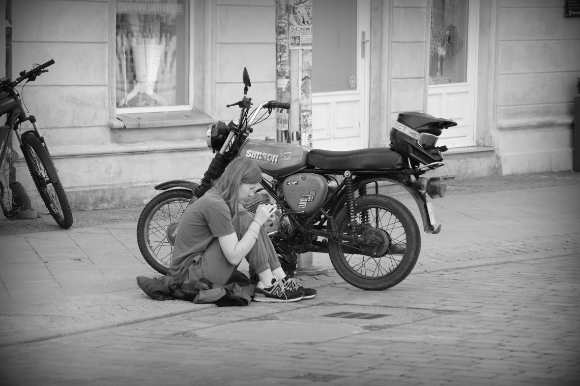 A monochrome photograph of a young person sitting on their jacket on the pavement checking their mobile phone. Behind them is a small Simson motorcycle with a driver's helmet propped on the seat. Copyright Urban Camera