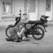 A monochrome photograph of a young person sitting on their jacket on the pavement checking their mobile phone. Behind them is a small Simson motorcycle with a driver's helmet propped on the seat. Copyright Urban Camera